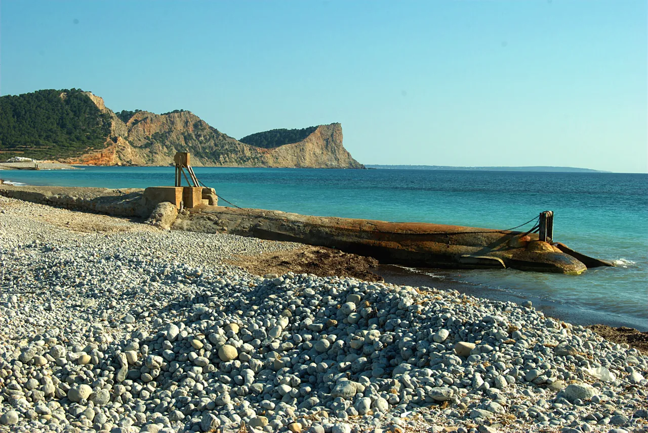 Strand aus weißen Steinen in Es Codolar mit dem Kap im Hintergrund